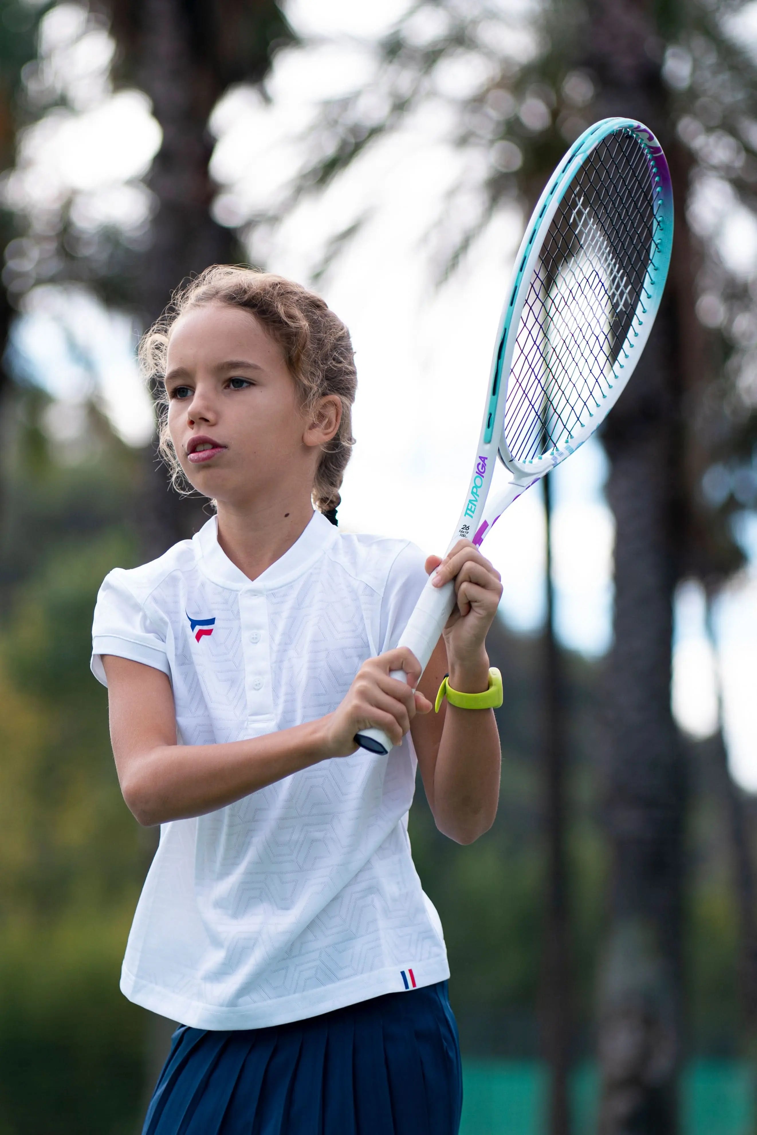 Young girl in white polo holding a Tecnifibre Tempo Iga 23 junior tennis racquet with purple and blue accents.