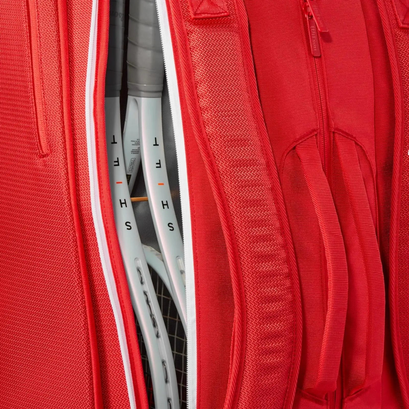 Close-up of a red Wilson Super Tour tennis bag with white zippers, showing multiple tennis racquets inside.