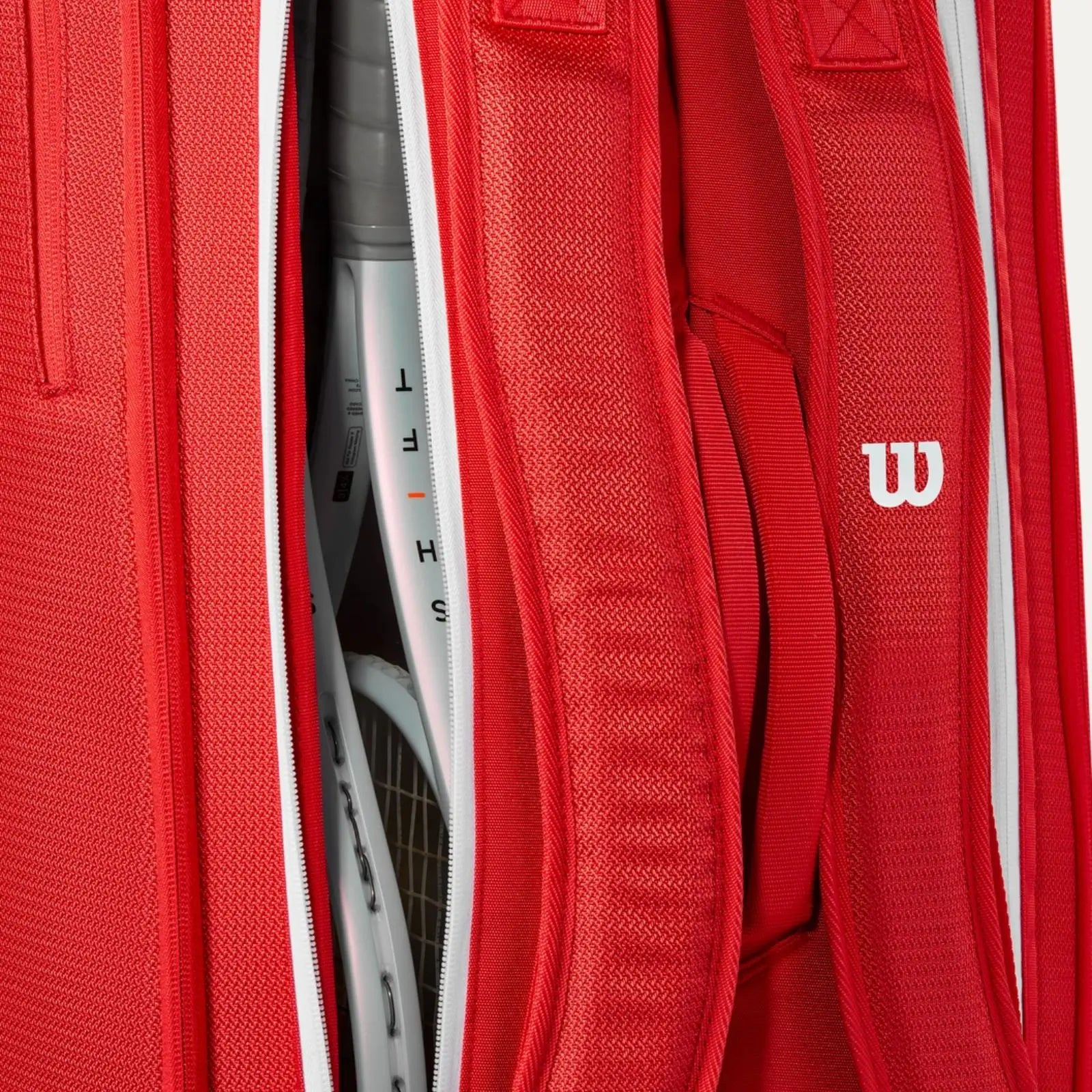 Red Wilson Super Tour tennis bag with white zippers, holding two white racquets, showing backpack straps and logo.