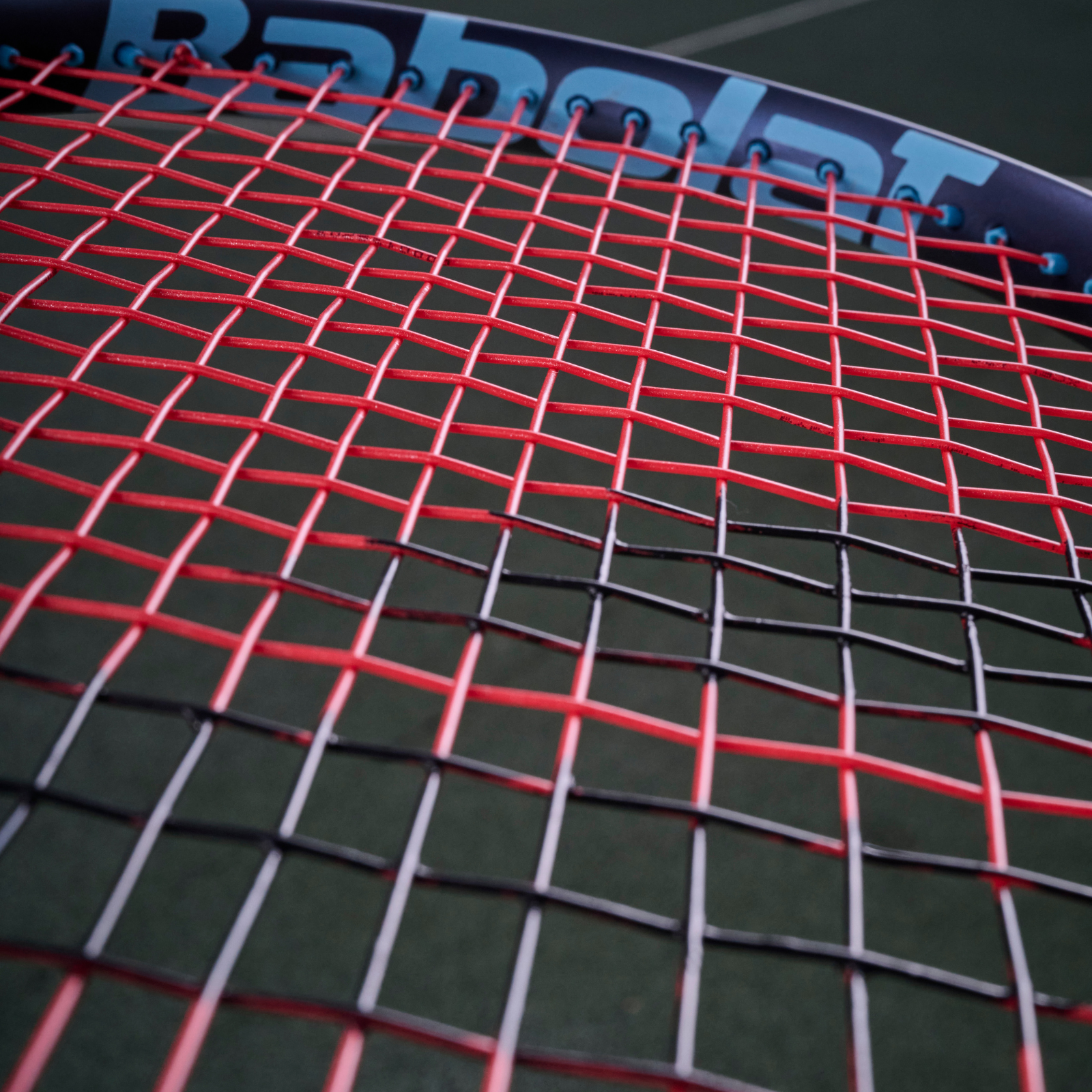 Close-up of a Babolat tennis racket strung with red and black XCEL 1.25mm multifilament string.