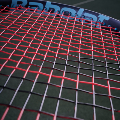Close-up of a Babolat tennis racket strung with red and black XCEL 1.25mm multifilament string.