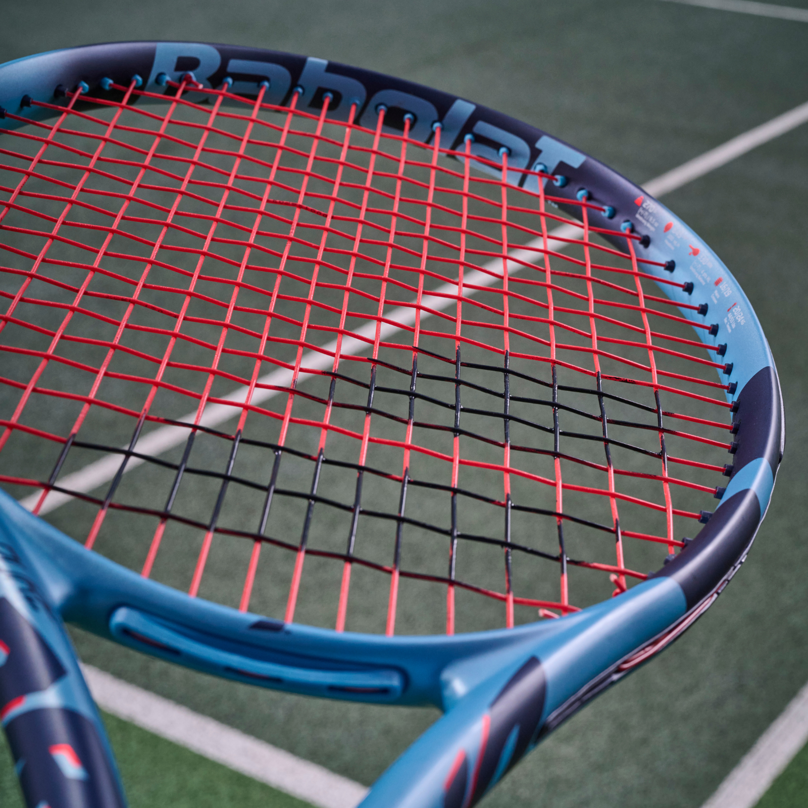 Babolat tennis racket with red XCEL 1.25 (17) multifilament strings, on a green court.