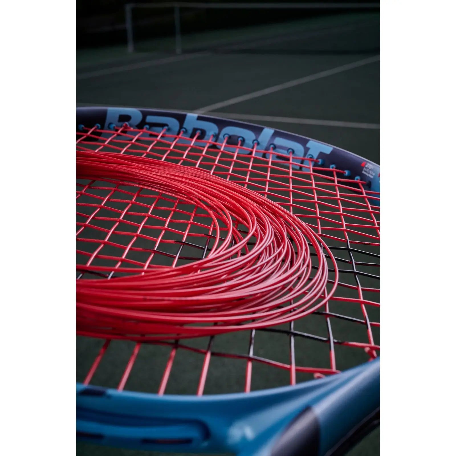 Coiled red Babolat XCEL tennis string on a blue tennis racket with a green court background.