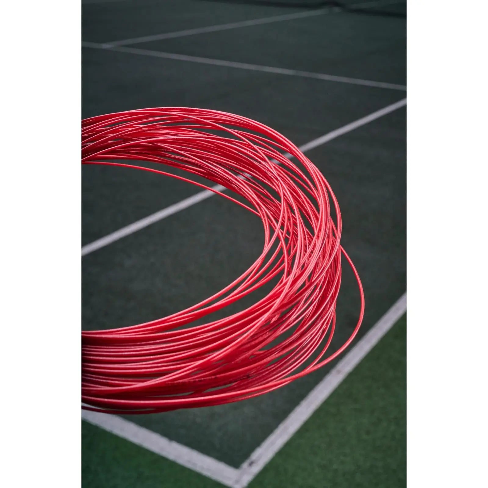Coiled red Babolat XCEL 1.30 (16) tennis string, with a blurred green tennis court in the background.
