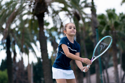 Young girl in navy shirt and white skirt holding a white and light blue Tecnifibre Tempo Iga 24 junior tennis racquet.