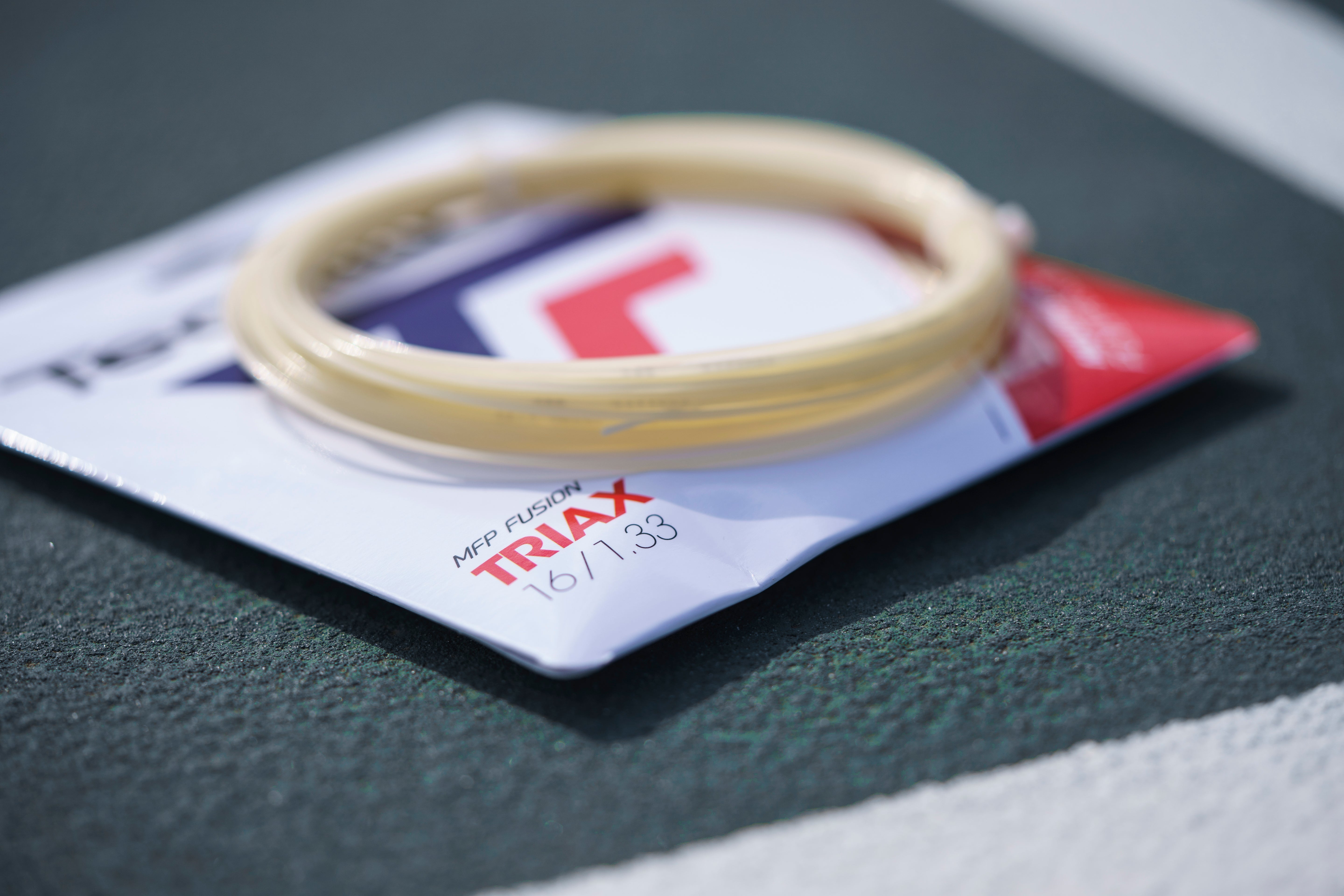 Natural Tecnifibre Triax 1.33 tennis string coil on its white and red package, resting on a green court.