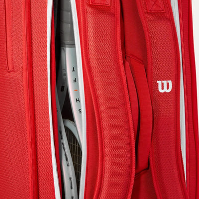 Red Wilson Super Tour tennis bag with white zippers, holding two white racquets, showing backpack straps and logo.